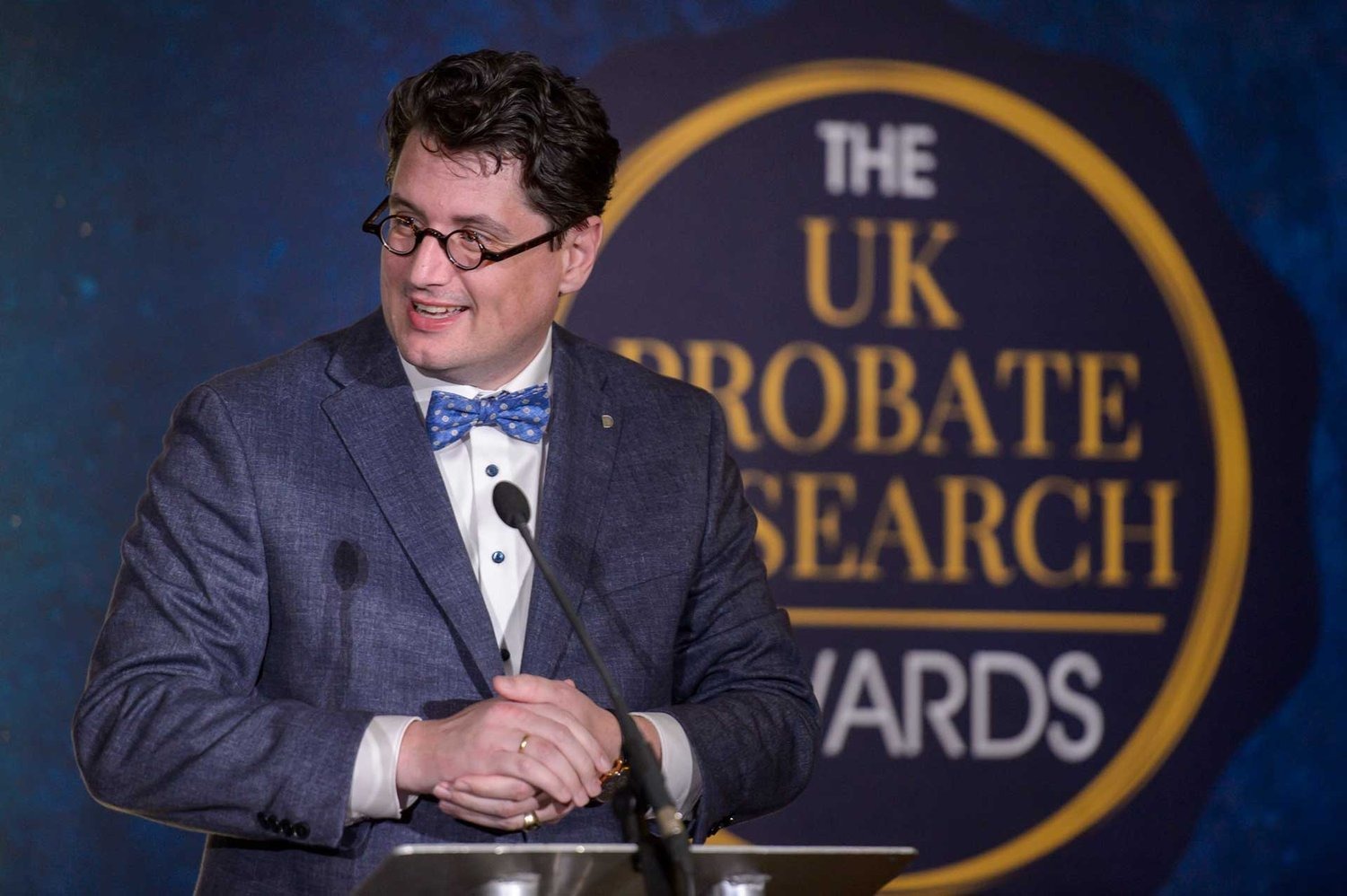 Klaas Zondervan, un homme en costume bleu et nœud papillon à pois, souriant depuis un podium, avec une pancarte « UK PROBATE SEARCH AWARDS » partiellement visible derrière lui.