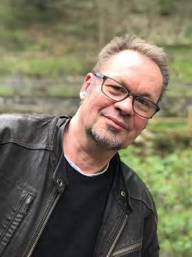 Portretfoto van Achim Bovelett. Een man met kort lichtbruin haar, een bril met een donker montuur en een sikje, gekleed in een donker leren jasje en een zwart shirt, kijkt met een lichte glimlach naar de camera.